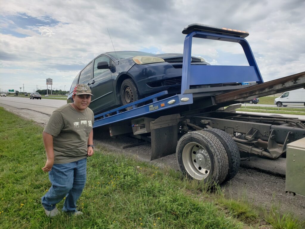 A boy smiling beside a car on a tow truck.
